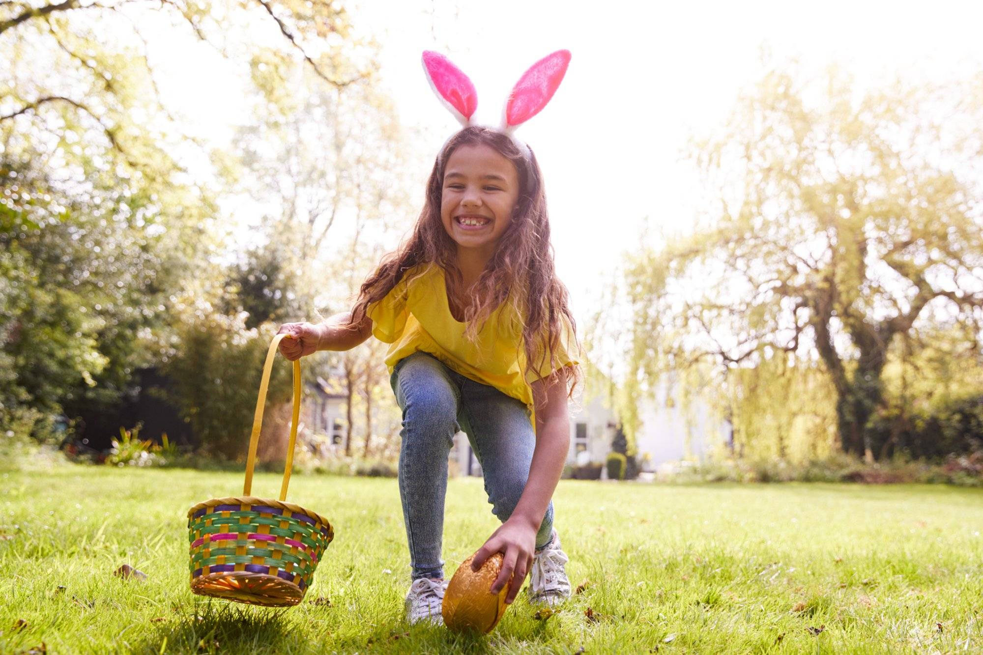 Portrait Of Girl Wearing Bunny Ears Finding Chocolate Egg On Easter Egg Hunt In Garden